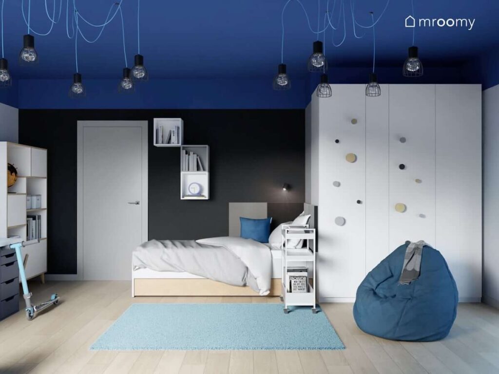 Niebiesko czarny pokój dla chłopca z dużą białą szafą ozdobioną wieszakami biało drewnianym łóżkiem szafką na kółkach i pufą sako