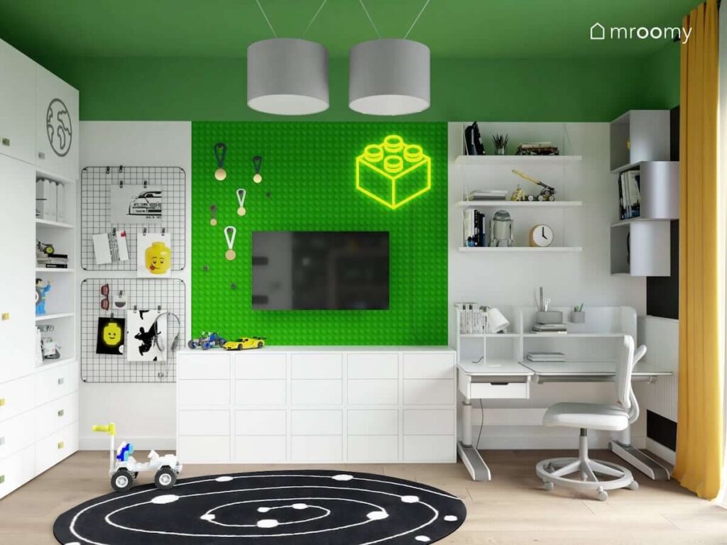 Strefa nauki z biurkiem z nadstawką oraz półkami ściennymi a także niska komoda i organizery ścienne oraz ściana oklejona zieloną naklejką we wzór klocka Lego w pokoju dla chłopca