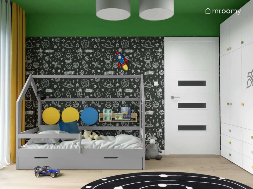Szare łóżko domek uzupełnione panelami ściennymi i półką na książki a za nim ściana pokryta kosmiczną tapetą w pokoju dla chłopca