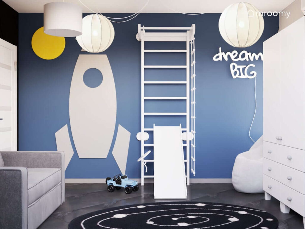 Niebieska ściana w pokoju kilkulatka a na niej drabinka gimnastyczna ze zjeżdżalnią oraz tablica kredowa w kształcie rakiety