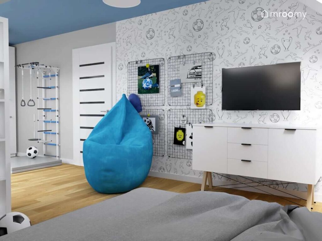 Ściana multimedialna pokryta piłkarska tapetą z telewizorem i komodą na drewnianych nogach a także organizery ścienne i niebieska pufa sako z biało niebieskim pokoju dla chłopca