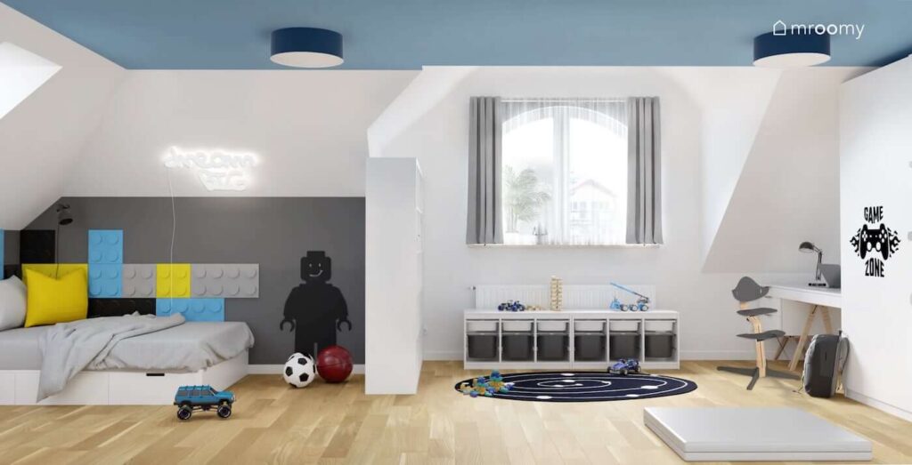 Biało szaro niebieski pokój dla chłopca w wieku szkolnym z niskim regałem z pojemnikami tablicą kredową w kształcie ludzika Lego i łóżkiem uzupełnionym kolorowymi panelami ściennymi
