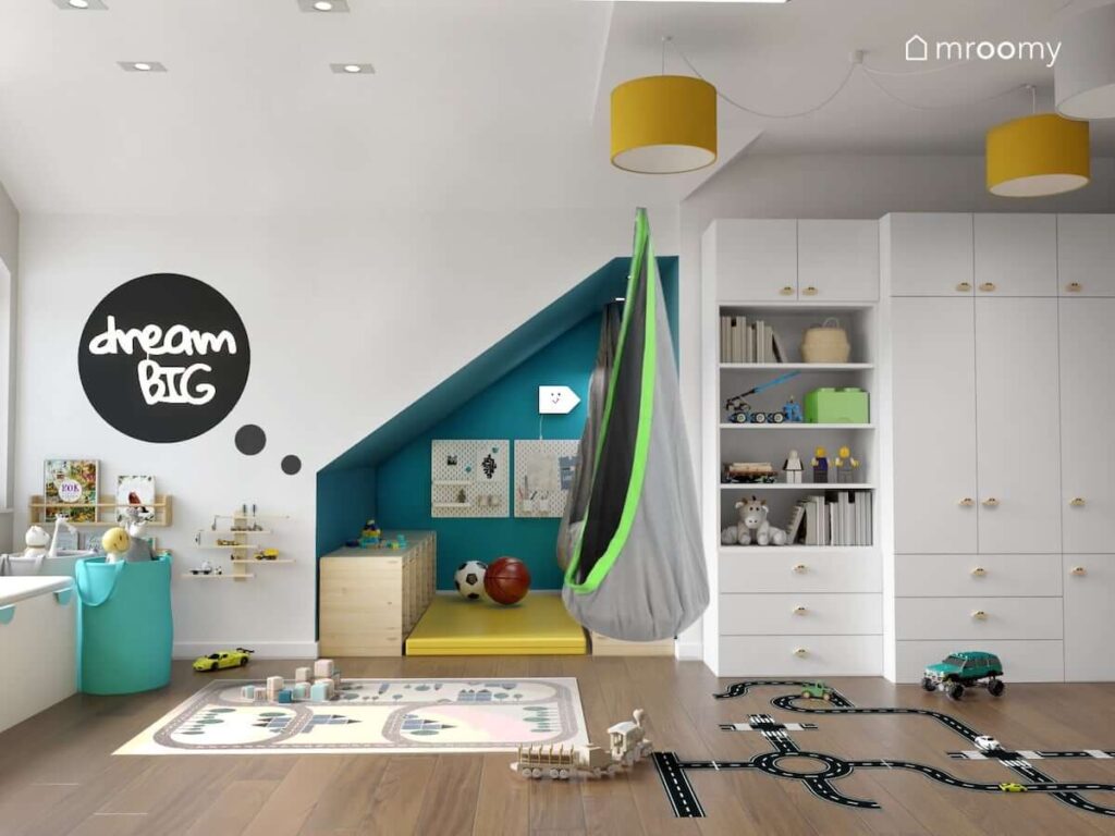 Jasny pokój dla małego chłopca z białą szafą z gałkami w kształcie samochodów a także huśtawką kokonem i turkusowym kącikiem z regałami na zabawki