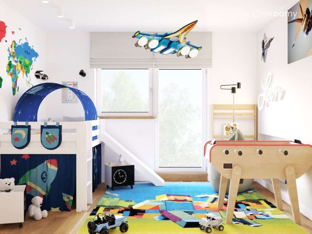 Jasny pokój dla chłopca w wieku szkolnym z białym łóżkiem ze zjeżdżalnią uzupełnionym dodatkami z motywami kosmicznymi a także ze stołem do gier kolorowym dywanem i lampą w kształcie samolotu