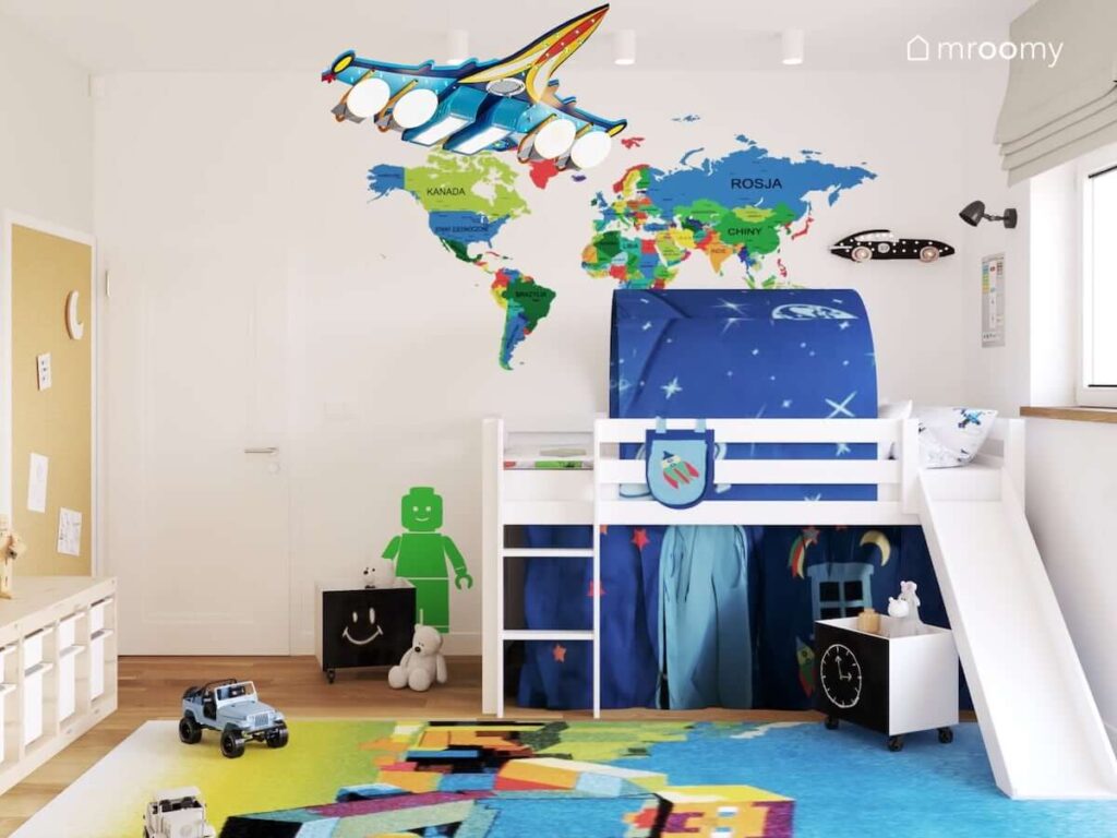 Białe łóżko ze zjeżdżalnią uzupełnione zasłonkami i dodatkami z motywami kosmicznymi oraz kolorowa mapa świata i zielona naklejka ścienna w kształcie ludzika Lego w pokoju dla chłopca