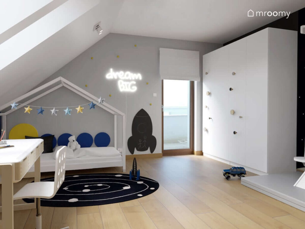 Biało szary kosmiczny pokój dla chłopca z łóżkiem domkiem ozdobionym girlandą gwiezdną i panelami ściennymi a także czarna tablica kredowa w kształcie rakiety ledon z kształcie napisu i duża biała szafa