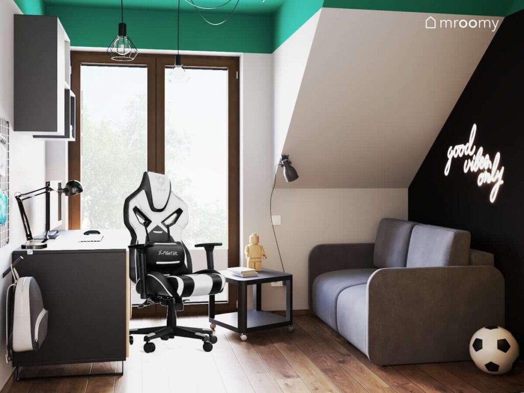 Szaro-czarny pokój dla nastolatka z zielonym sufitem rozkładaną sofą oraz czarno drewnianym biurkiem z fotelem gamingowym