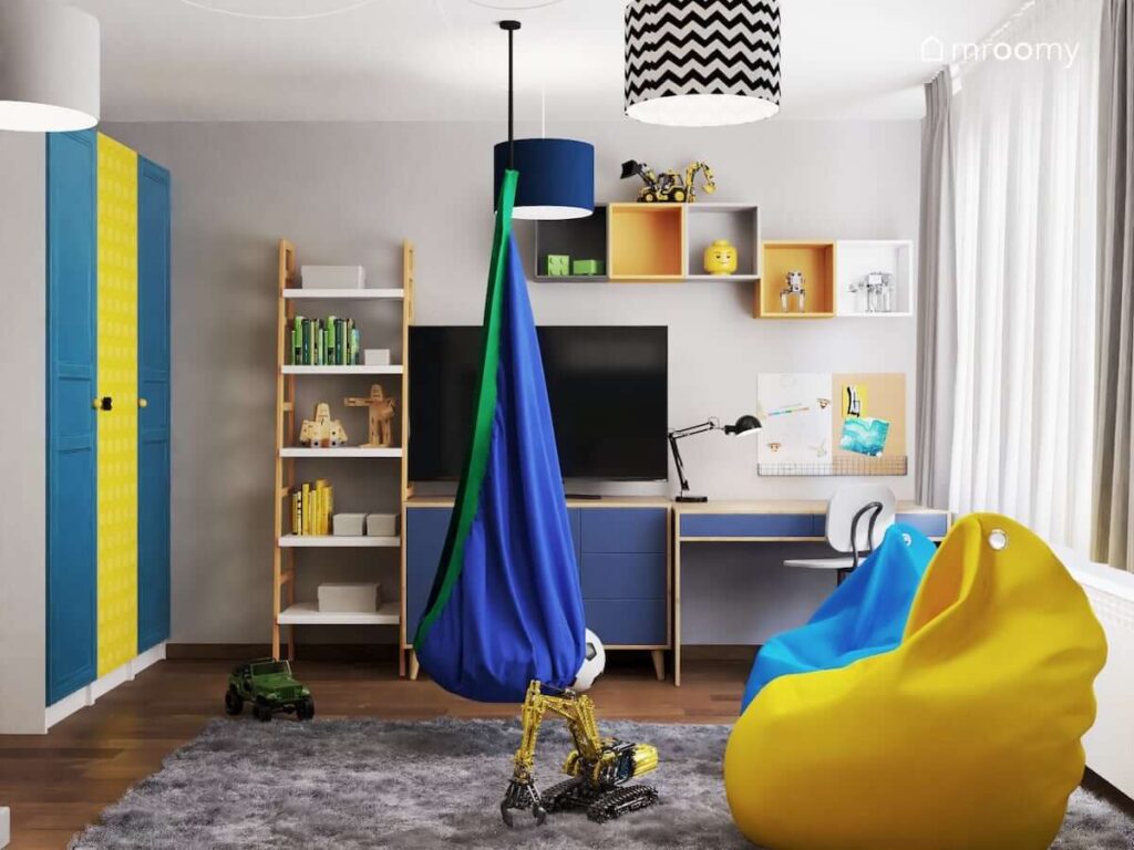 Strefa nauki w pokoju dla chłopca w wieku szkolnym a w nim niebiesko drewniane biurko niebiesko drewniana komoda telewizor regał w kształcie drabinki oraz szafki ścienne w różnych kolorach
