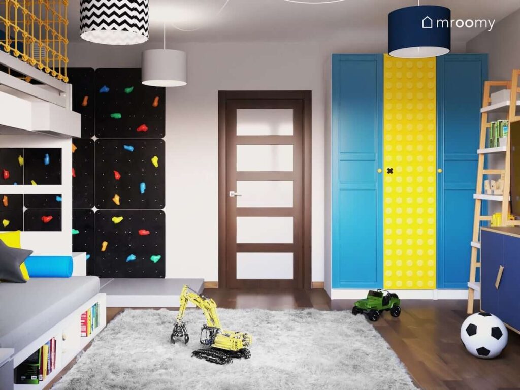 Czarna ścianka wspinaczkowa z kolorowymi uchwytami a także niebieska szafa z żółtą naklejką we wzór klocka Lego i szary futerkowy dywan w pokoju dla ucznia