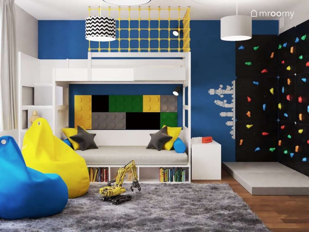 Biało szare łóżko piętrowe z kolorowymi poduszkami oraz panelami ściennymi w kształcie klocków i żółtą siatką zabezpieczającą a także czarna ścianka wspinaczkowa z kolorowymi uchwytami w pokoju dla chłopca