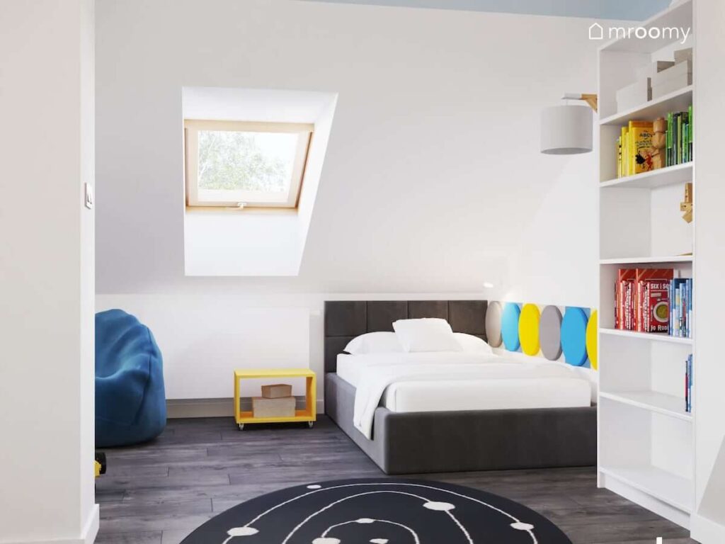 Strefa spania w poddaszowym pokoju dla chłopca a w niej szare łóżko uzupełnione kolorowymi panelami ściennymi i żółty stolik nocny na kółkach