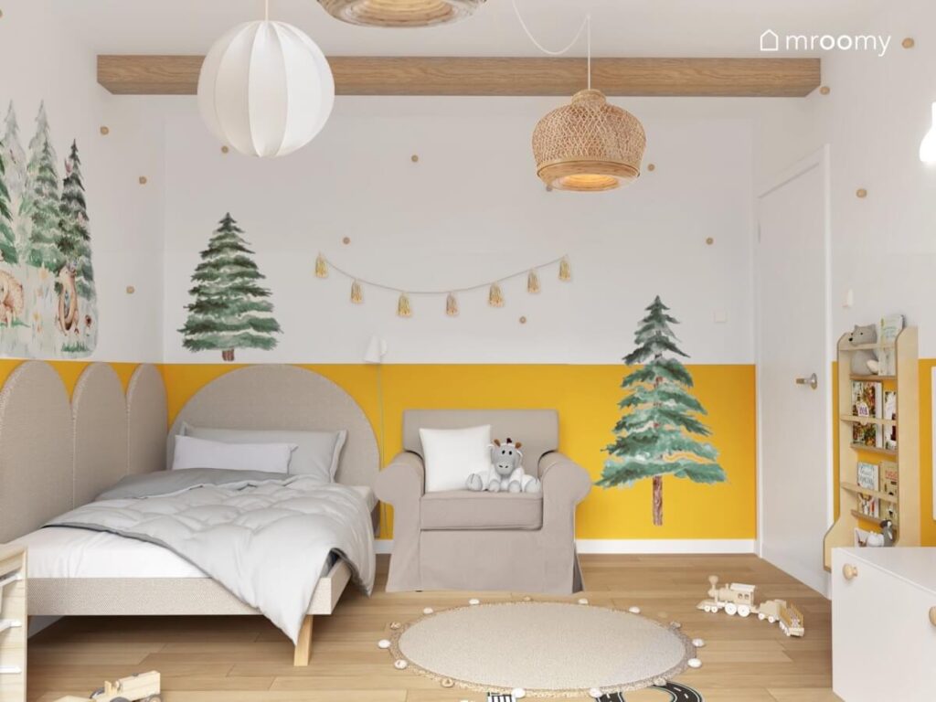 Strefa spania w biało żółtym pokoju dla chłopca a w nim szare tapicerowane łóżko szary fotel oraz ściana ozdobiona girlandą z pomponów i naklejkami w kształcie choinek