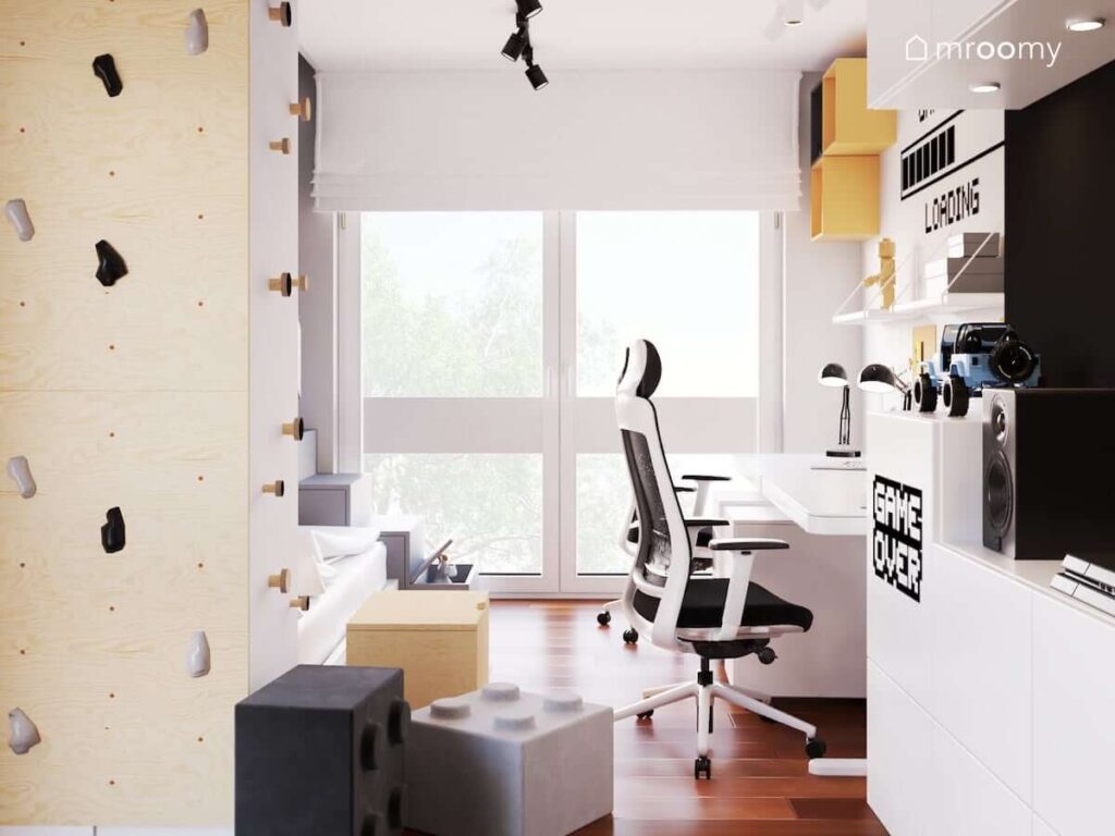 Dwa białe biurka a także ścianka wspinaczkowa i szare pufki w kształcie klocków w pokoju dwóch braci