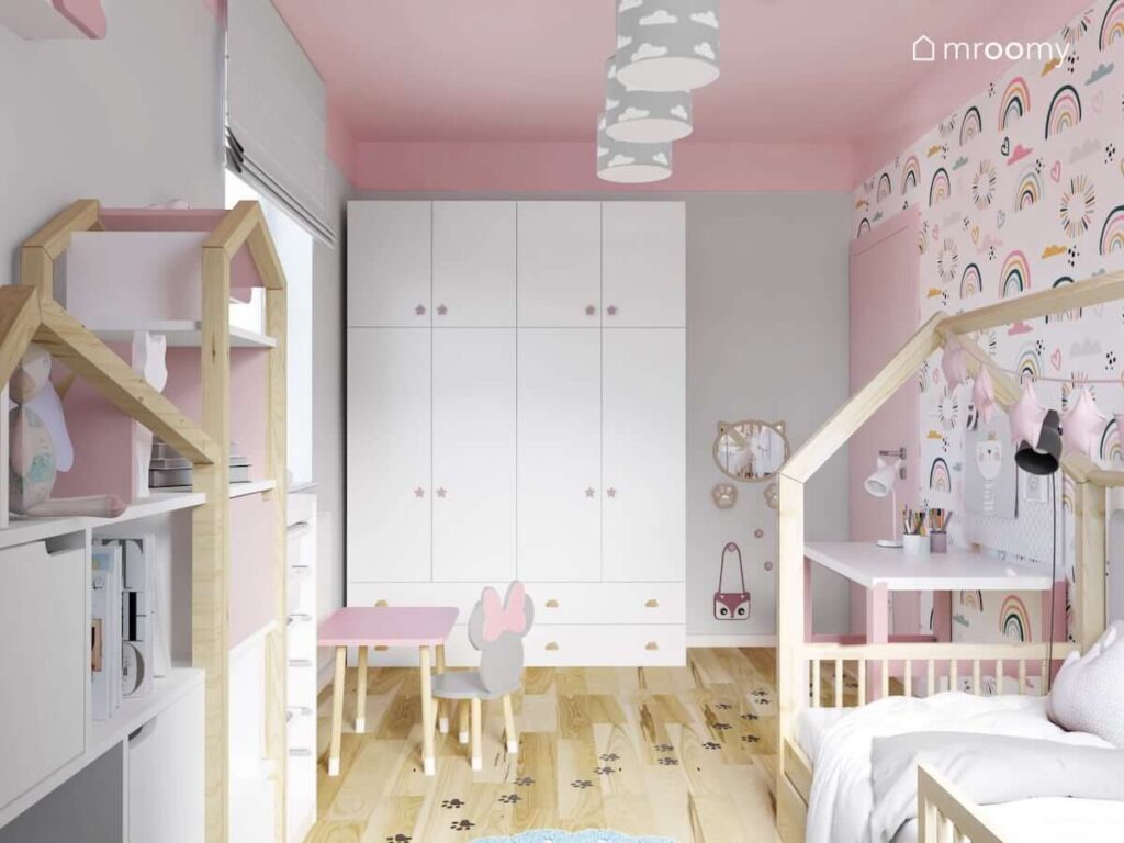 Biała szafa uzupełniona gałkami w kształcie gwiazd a także lustro w kształcie kotka i różowy stolik na drewnianych nogach z krzesełkiem z oparciem w kształcie Myszkie Minnie w pokoju dla dwóch dziewczynek