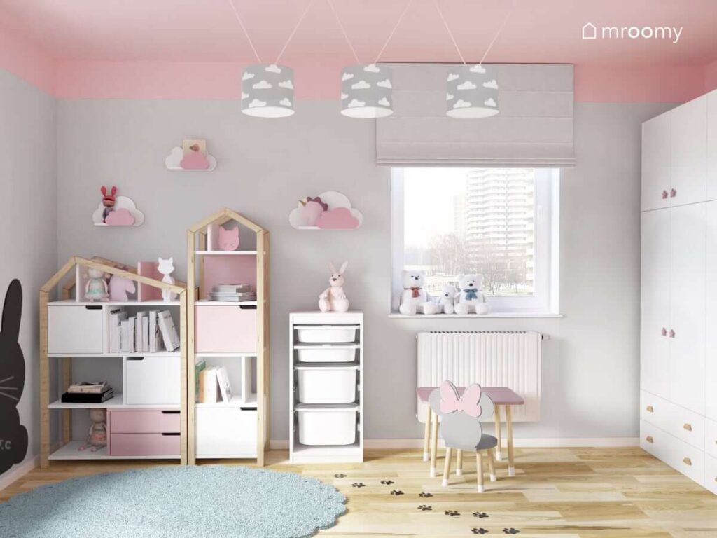 Biało drewniano różowe regały w kształcie domków półki w kształcie chmurek oraz regał z pojemnikami na zabawki a na suficie lampy z abażurami w chmurki w szaro różowym pokoju dwóch sióstr