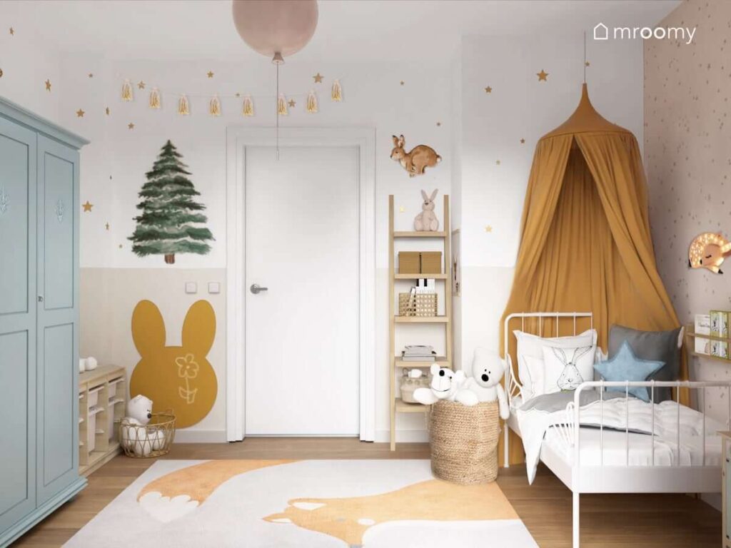 Białe łóżko ozdobione karmelowym baldachimem a także drewniany regał w kształcie drabinki tablica kredowa w kształcie głowy królika i dywan z lisem w pokoju dla dziewczynki