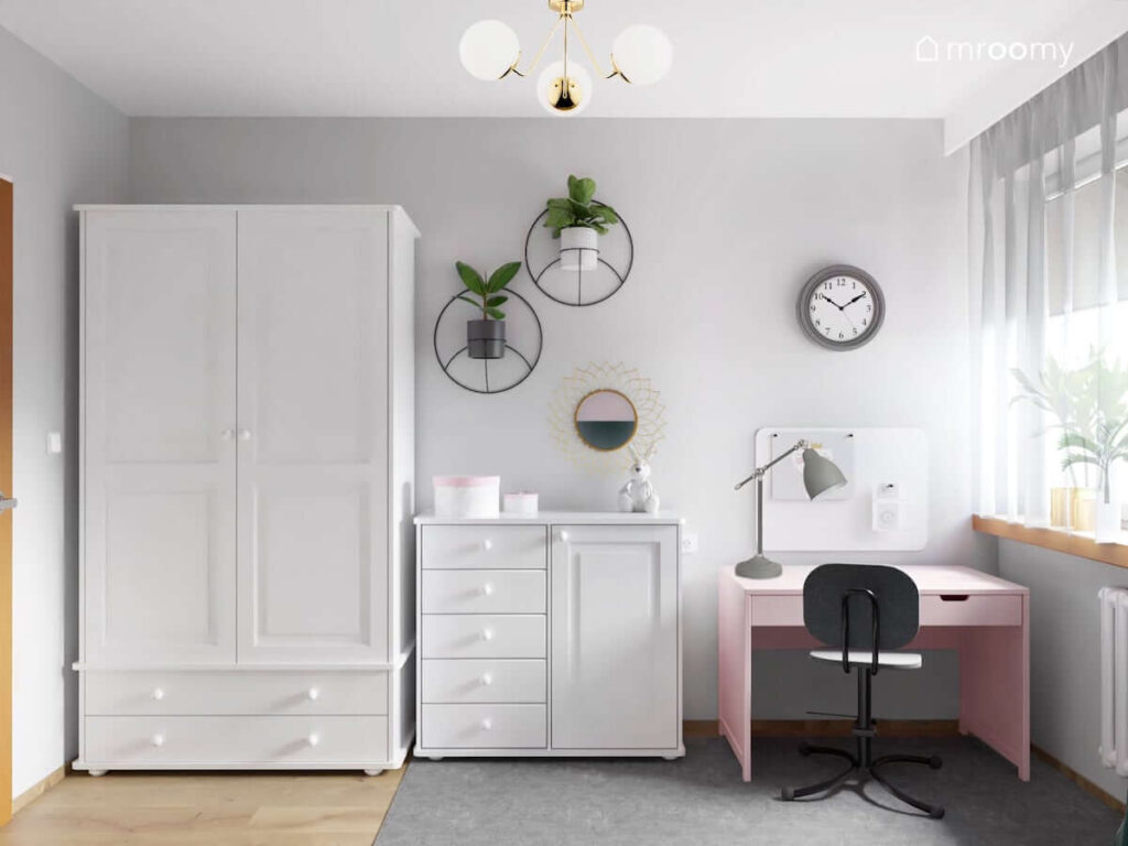 Biała szafa dwudrzwiowa komoda z szufladami różowe biurko a nad nimi zegar kwietniki oraz ozdobne lustro w pokoju dla dziewczynki w wieku przedszkolnym