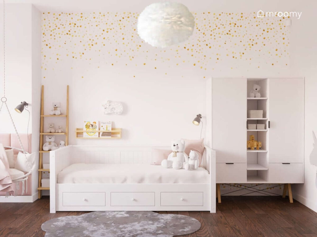 Pokój dziewczynki w wieku szkolnym z białym łóżkiem szafą na drewnianych nogach drewnianym regałem w kształcie drabinki oraz ze ścianą ozdobioną tapetą w złote kropki