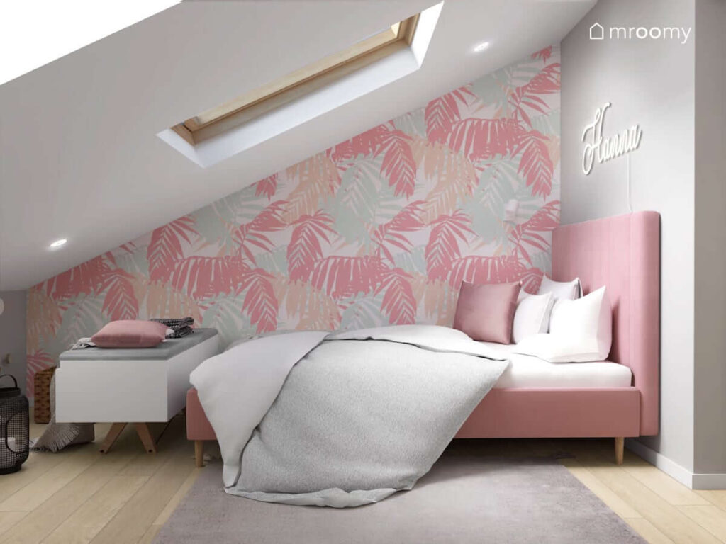 Strefa spania w poddaszowym pokoju dla nastolatki z różowym łóżkiem z wysokim zagłówkiem oraz ścianami ozdobionymi tapetą w liście i ledonem w kształcie imienia
