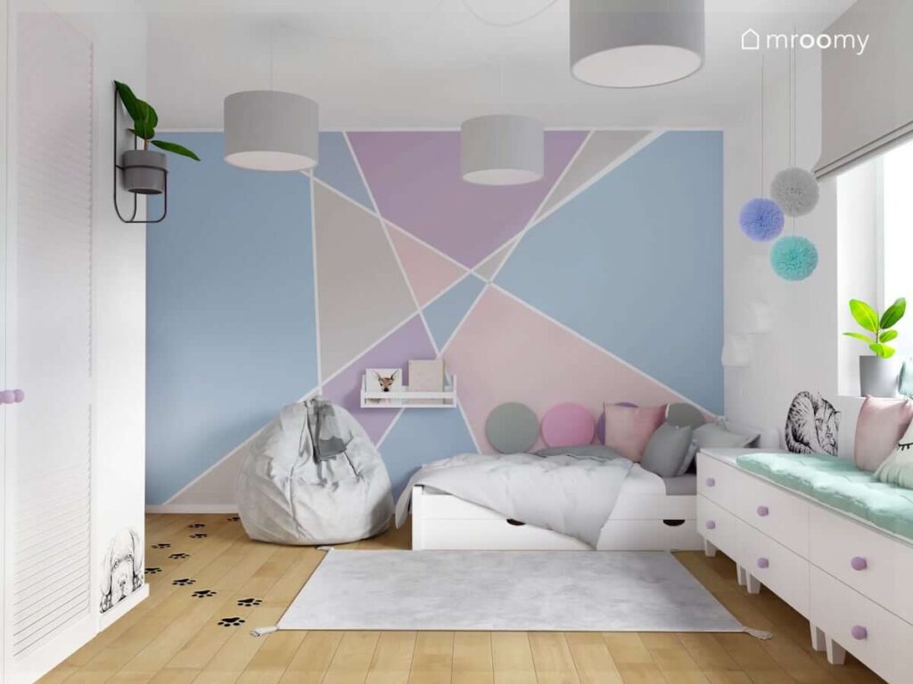 Strefa spania w pokoju dla dziewczynki z białym łóżkiem uzupełnionym panelami ściennymi pufą sako oraz ścianą ozdobioną geometrycznym malunkiem w kolorach pastelowych