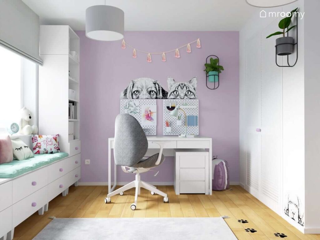 Fioletowa strefa nauki w pokoju dla dziewczynki z białym biurkiem szarym krzesłem oraz organizerami kwietnikiem girlandą i naklejkami z psem i kotem na ścianie