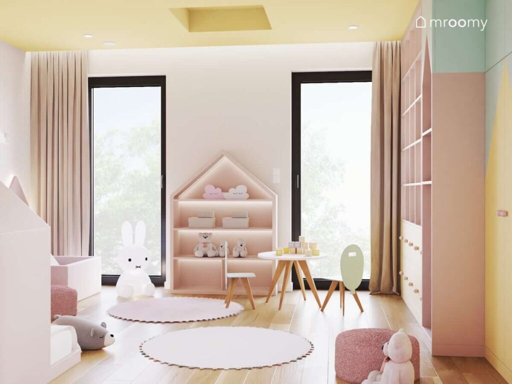 Regał w kształcie domku a także stolik z krzesełkami z oparciami w kształcie balonów i dwa okrągłe dywany w różowo beżowo żółtym pokoju dla dziewczynki