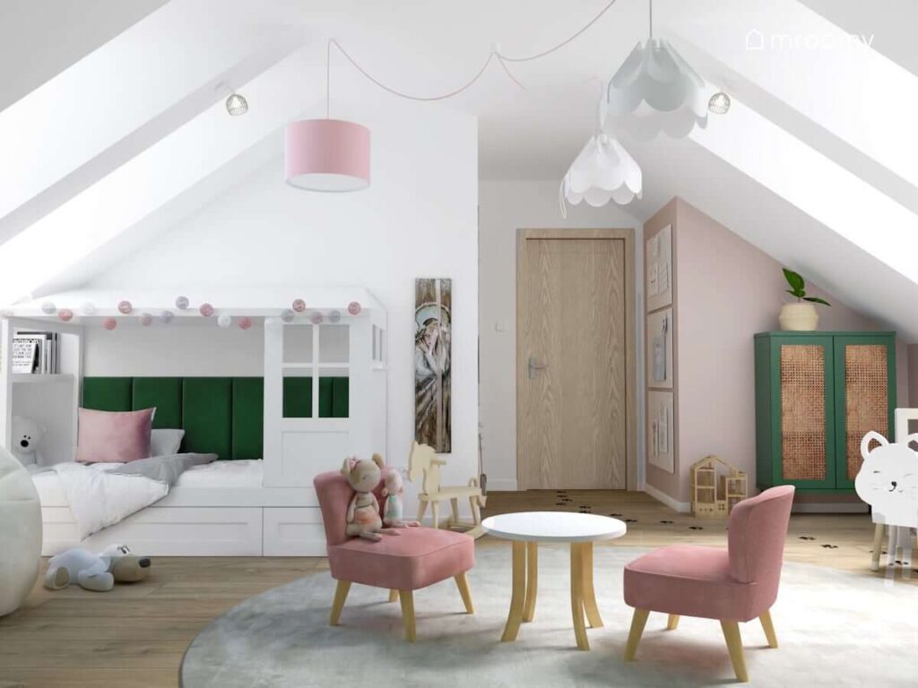 Jasny poddaszowy pokój dla dziewczynki z białym łóżkiem domkiem uzupełnionym zielonymi panelami ściennymi oraz girlandą cotton balls a także z okrągłym stolikiem z różowymi fotelikami