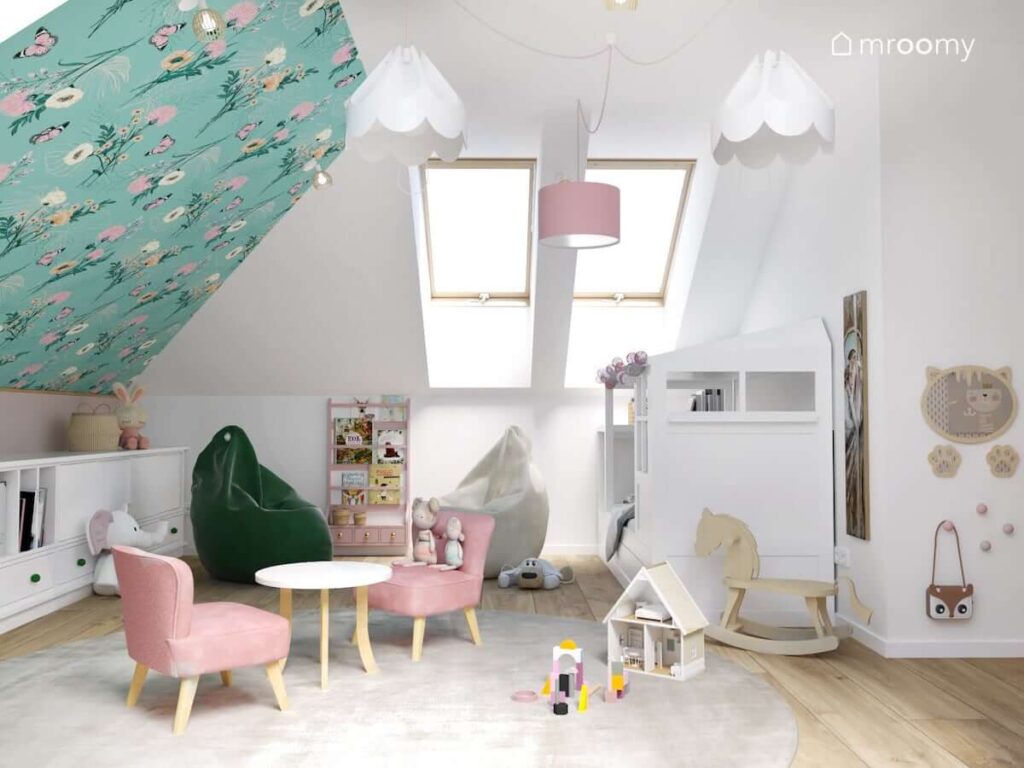 Biały poddaszowy pokój dla dziewczynki ze skosem ozdobionym tapetą w kwiaty a także z białym łóżkiem białą i zieloną pufą sako różową biblioteczką i lustrem w kształcie kotka