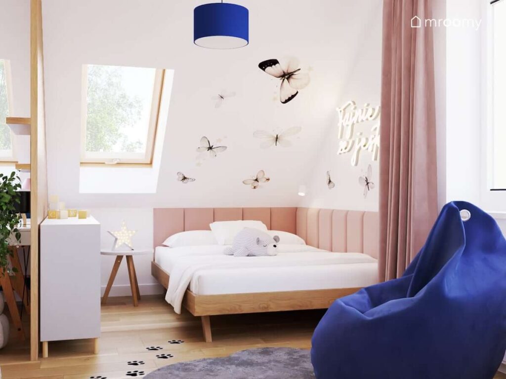 Strefa spania z drewnianym łóżkiem uzupełnionym panelami ściennymi a także skos ozdobiony naklejkami w kształcie motyli i ledon w kształcie napisu w pokoju dla dziewczynki