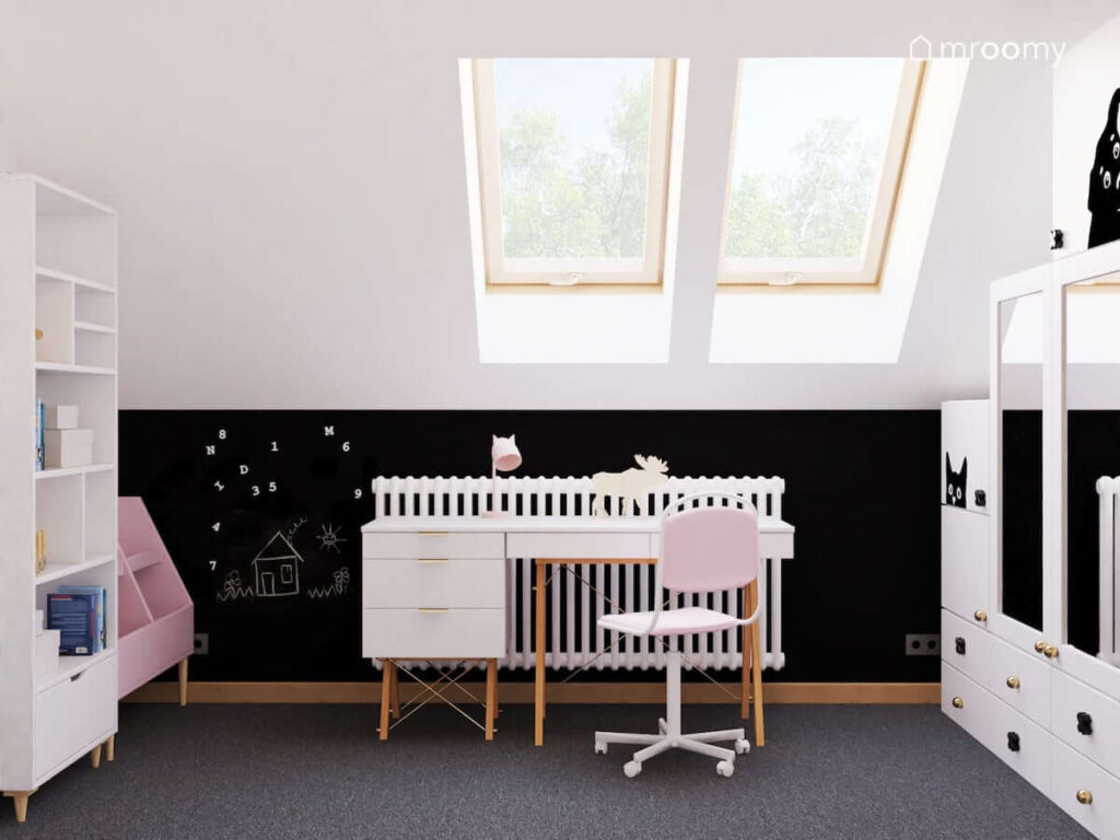 Strefa nauki z białym biurkiem na drewnianych nogach oraz kontenerkiem i ścianą pomalowaną farbą tablicową w biało czarnym poddaszowym pokoju dla dziewczynki