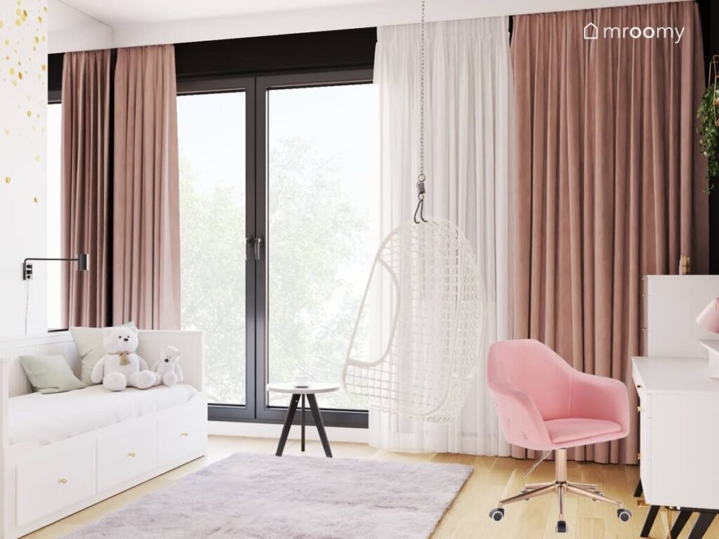 Pokój dla nastolatki z dużym oknem uzupełnionym firanką i zasłonami a także z białym łóżkiem fotelem wiszącym różowym krzesłem biurowym i szarym dywanem
