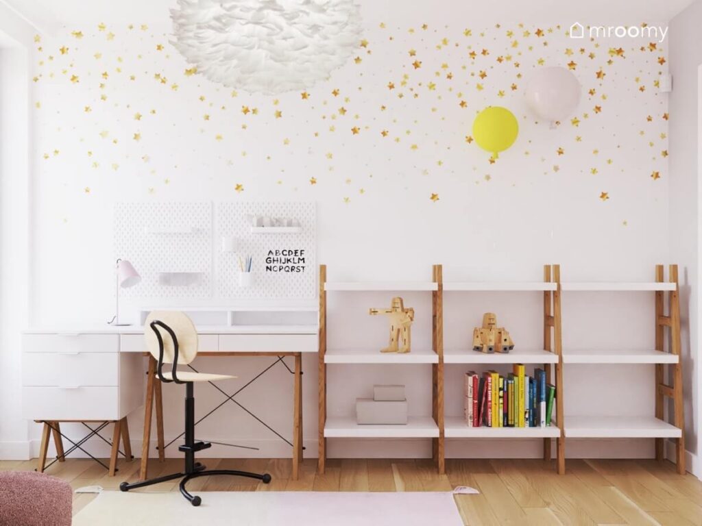 Strefa nauki w pokoju dla dziewczynki z biurkiem na drewnianych nogach niskim regałem w kształcie drabinki organizerami ściennymi oraz ściana pokrytą tapetą w złote gwiazdki i z kinkietami w kształcie balonów