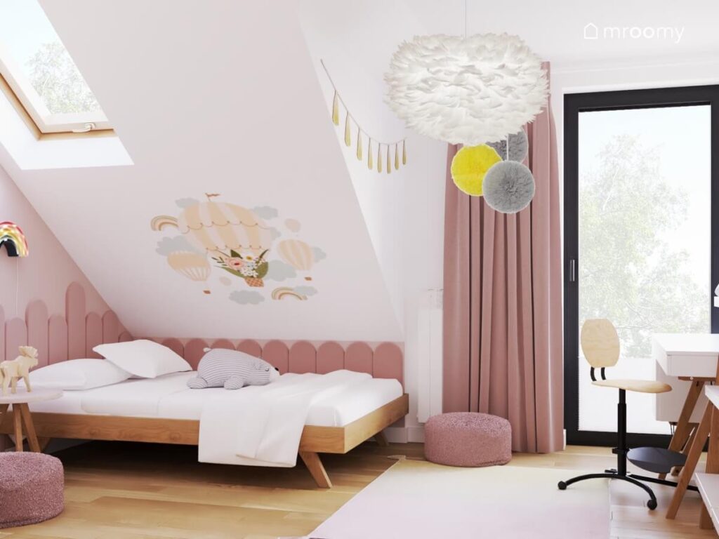Strefa spania w pokoju dla dziewczynki z drewnianym łóżkiem uzupełnionym różowymi panelami ściennymi oraz z naklejką w kształcie balonów powietrznych i chmurek a u sufitu lampa z ptasich piór
