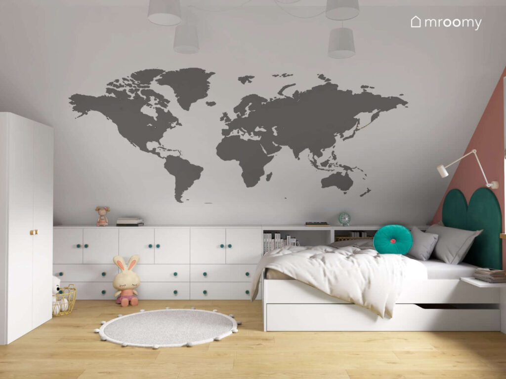 Strefa spania w poddaszowym pokoju dla dziewczynki z białym łóżkiem oraz niską zabudową meblową a na ścianie tapeta z mapą świata