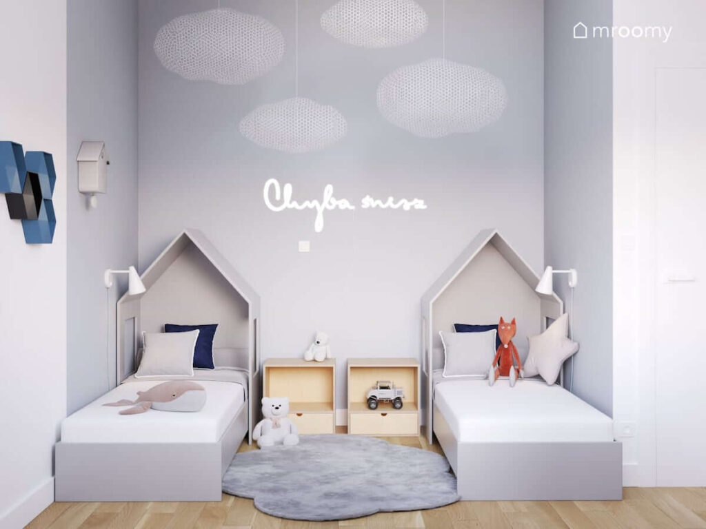 Strefa spania w pokoju dla rodzeństwa z łózkami w kształcie domków dywanem w kształcie chmurki oraz z ledonem w kształcie napisu i wiszącymi dekoracyjnymi chmurkami