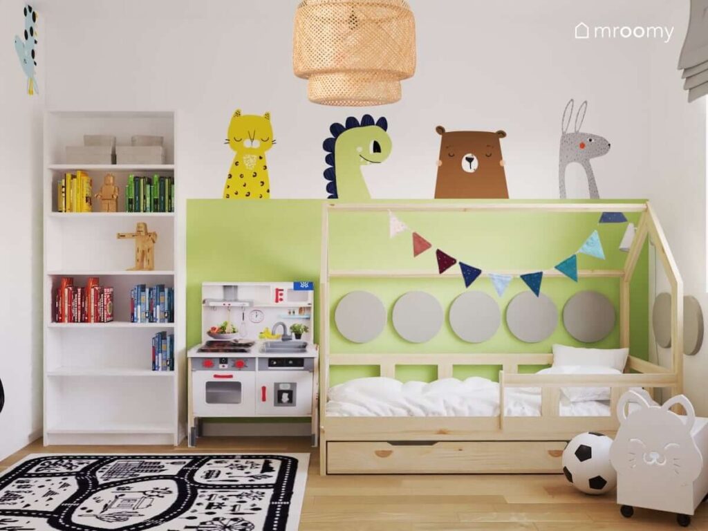 Drewniane łóżko w kształcie domku kuchenka dla dzieci oraz biały regał a na zielonej ścianie naklejki w kształcie zwierzątek w pokoju dla chłopca i dziewczynki