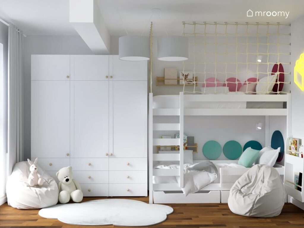 Duża biała szafa oraz białe łóżko piętrowe uzupełnione panelami ściennymi w różnych kolorach a także dywanik w kształcie chmurki w pokoju dla rodzeństwa