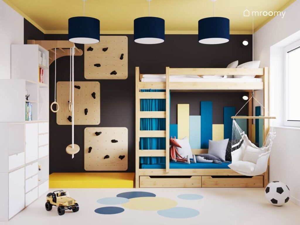 Drewniane łóżko piętrowe uzupełnione kolorowymi panelami ściennymi oraz zasłonką a także ścianka wspinaczkowa i fotel wiszący w czarno żółtym pokoju dla chłopca