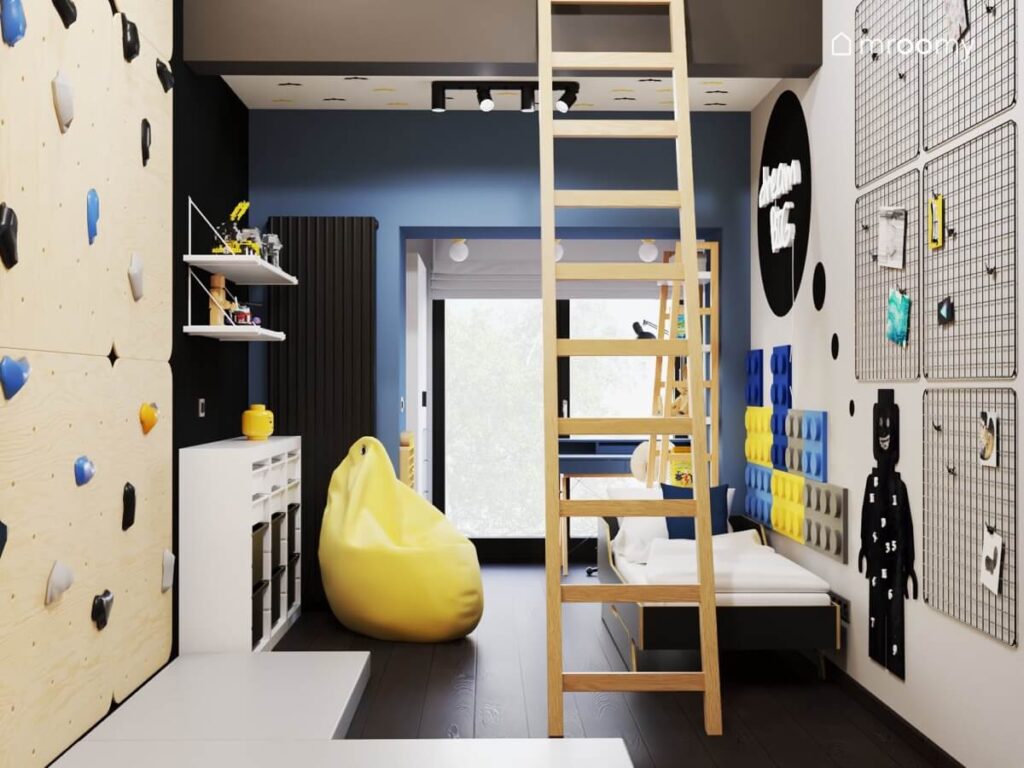 Biało niebiesko czarny pokój dla chłopca a w nim ściana wspinaczkowa żółta pufa biały regał z pojemnikami na zabawki oraz drabina prowadząca na antresolę