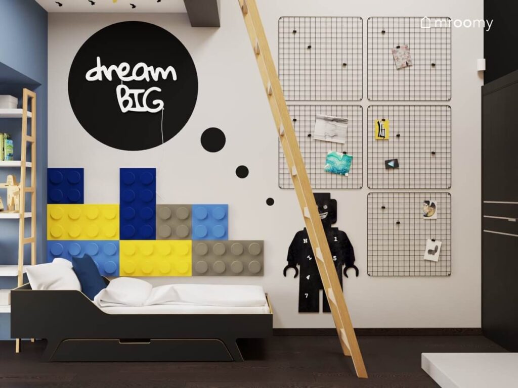 Czarne łóżko uzupełnione kolorowymi panelami ściennymi w kształcie klocków a nad nim ledon w kształcie napisu a obok tablica kredowa w kształcie ludzika Lego oraz organizery w pokoju dla chłopca