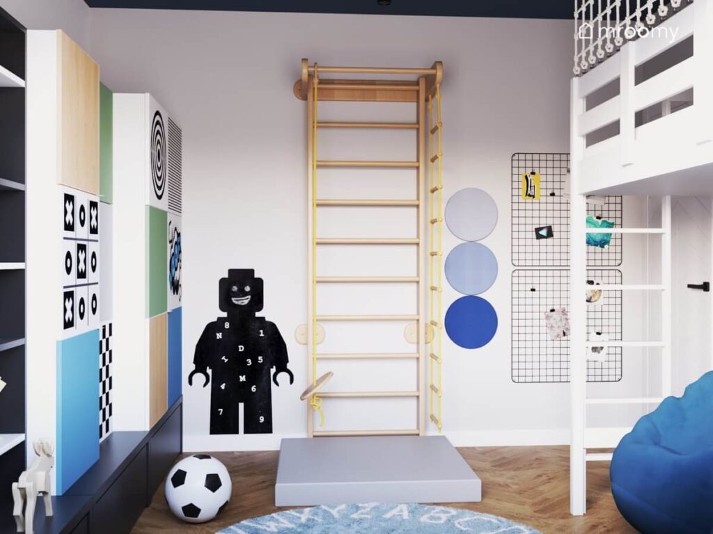 Drewniana drabinka gimnastyczna z materacem czarna tablica kredowa w kształcie ludzika Lego a także niebieski dywan z alfabetem i panele ścienne w różnych odcieniach niebieskiego w pokoju dla chłopca
