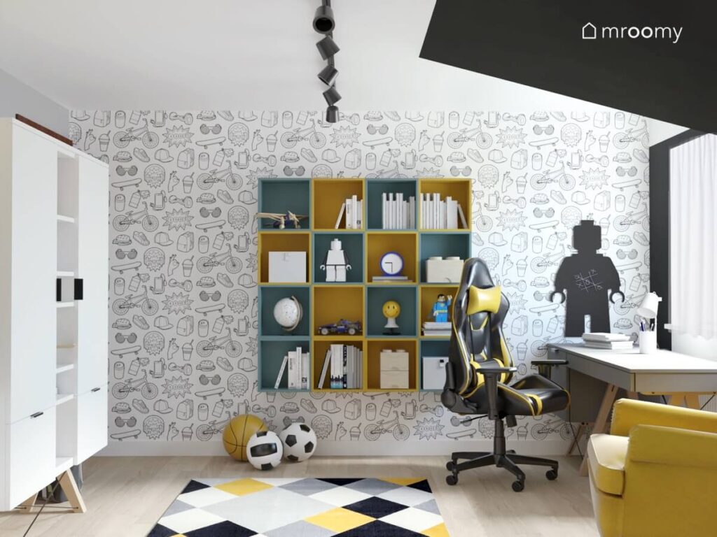 Ściana w pokoju dla chłopca ozdobiona młodzieżową tapetą a na niej tablica kredowa w kształcie ludzika Lego oraz żółte i szaroturkusowe szafki ścienne