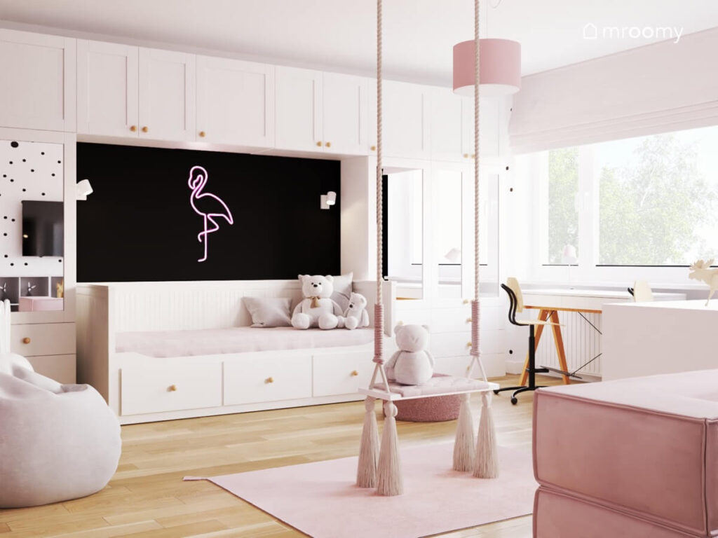 Huśtawka pokojowa z frędzlami oraz biała leżanka z szufladami a nad nią białe meble modułowe i ledon w kształcie flaminga w pokoju dla dziewczynki