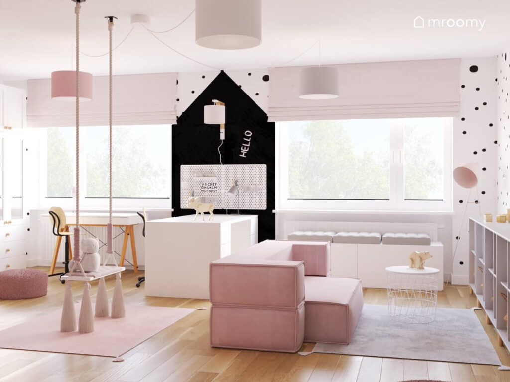Biało różowy pokój dla dziewczynki z huśtawką z frędzlami różową sofą białym siedziskiem z poduszkami stołem z miejscem do przechowywania i fragmentem ściany pomalowanym czarną farbą tablicową