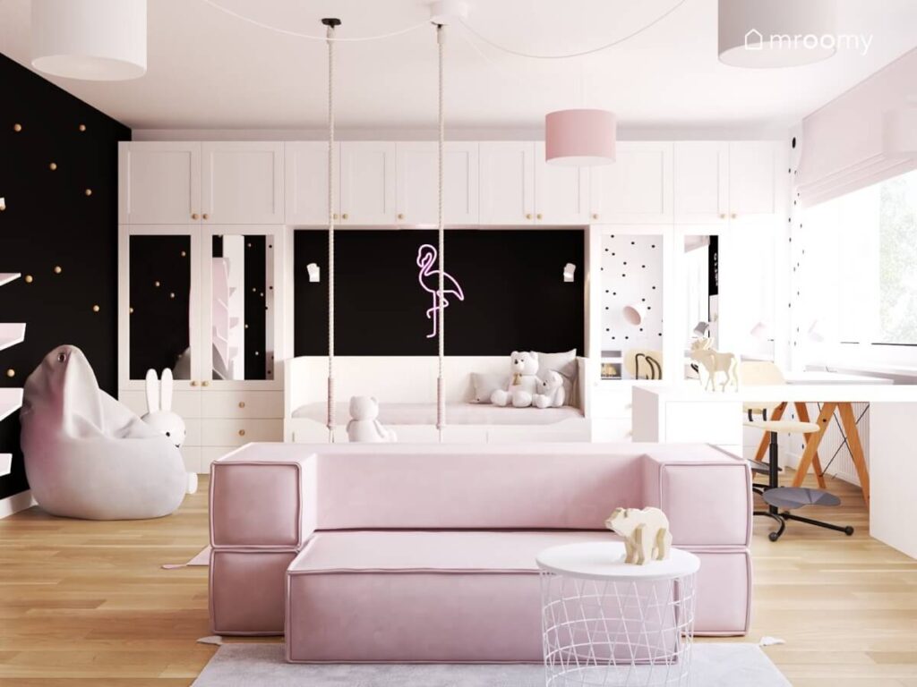 Białe meble modułowe różowa sofa pufa sako oraz ledon w kształcie flaminga na czarnej ścianie w pokoju dla dziewczynki