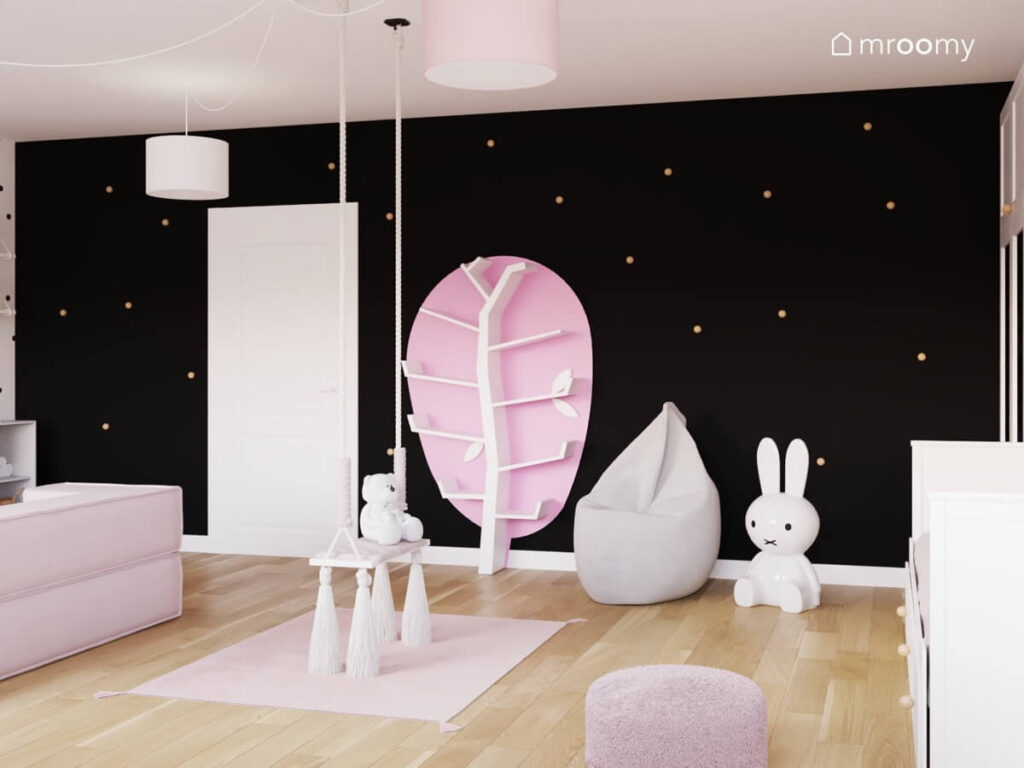 Czarna ściana w pokoju dla dziewczynki ozdobiona złotymi gałkami do mebli a na niej również różowo biały regał w kształcie drzewa pufa sako i lampa w kształcie królika