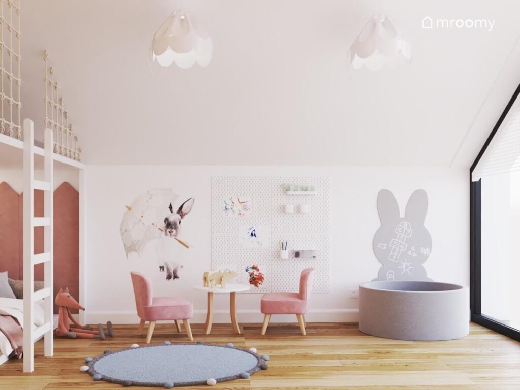 Biały pokój dla dziewczynki a w nim ściana z naklejką w kształcie królika organizerami i tablicą w kształcie królika a także basen z kulkami i stolik z welurowymi krzesełkami