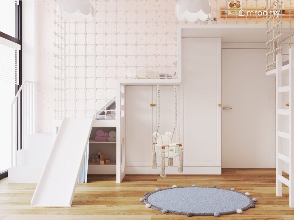 Biały pokój dla dziewczynki z antresolą zabezpieczoną siatką a pod nią szafy a oprócz tego huśtawka wisząca i niebieski dywan z pomponami