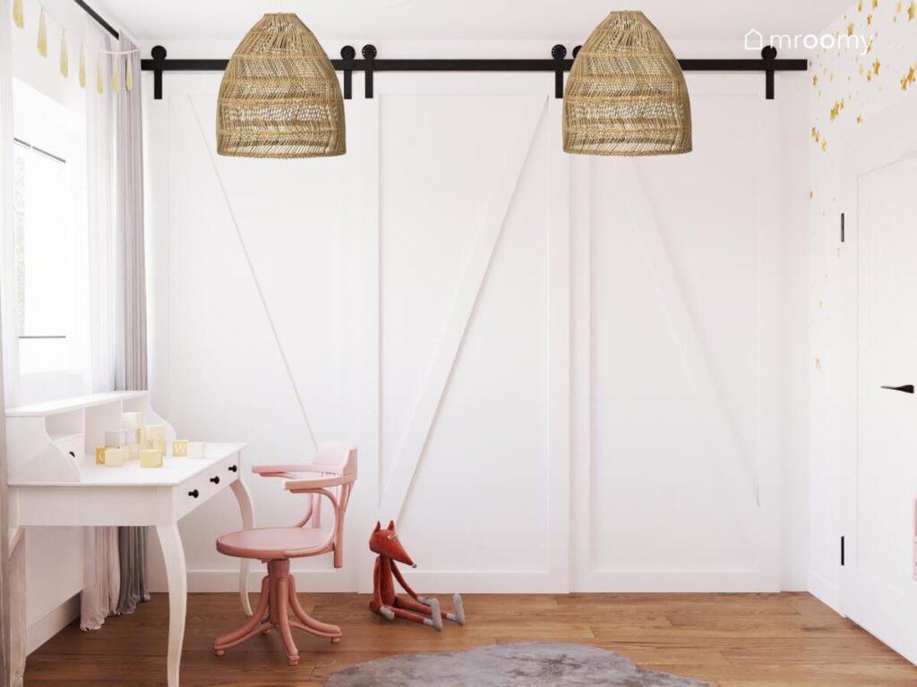 Duża biała szafa a także rattanowe lampy sufitowe i białe biurko z różowym krzesłem w pokoju dziewczynki