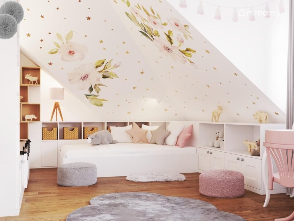 Biały pokój dla dziewczynki ze skosem ozdobionym naklejkami w kształcie magnolii i złotych gwiazdek a także białym łóżkiem i niską zabudową meblową